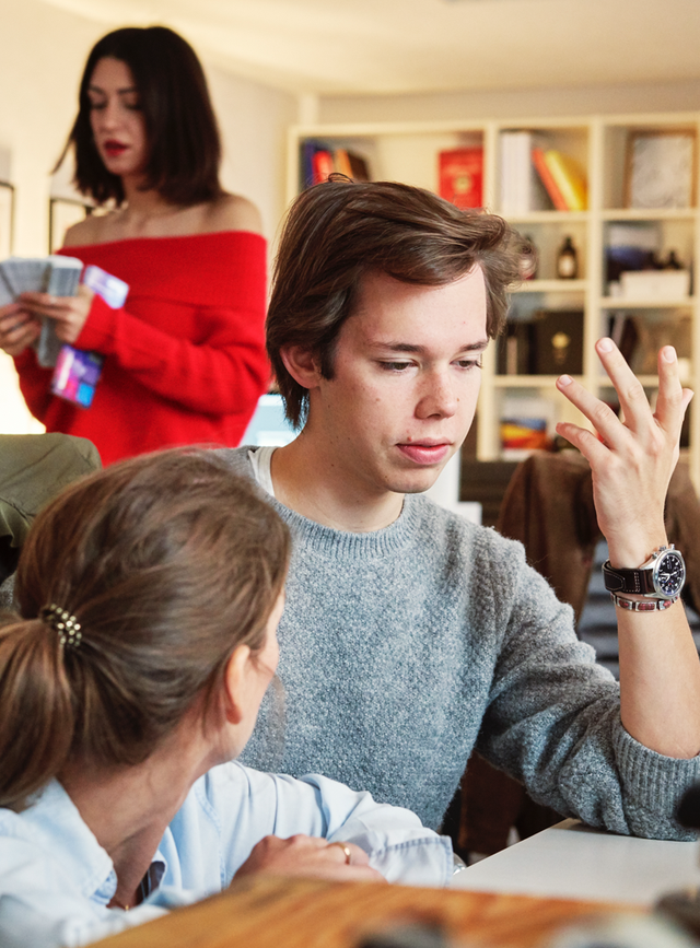 Ein Mann und eine Frau arbeiten zusammen an einem Schreibtisch und unterhalten sich. Im Hintergrund ist eine weitere Frau zu sehen, die nicht erkennbare Objekte in der Hand hält.