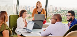 Image of 3 female and 2 male professionals sitting around a table with their laptops in a conference room, engaged in conversation.