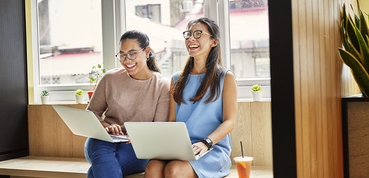 Two people laugh joyfully while working side by side with their laptops on their knees.