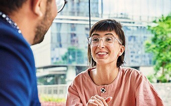 Photograph of a woman wearing a pink short wearing glasses with short brown hair talking to a man who is partially off the screen.