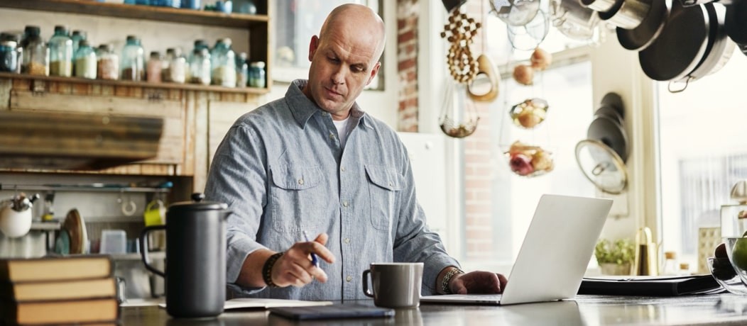 Caucasian man standing up in a kitchen with a notebook and a pen and notepad in front of him.