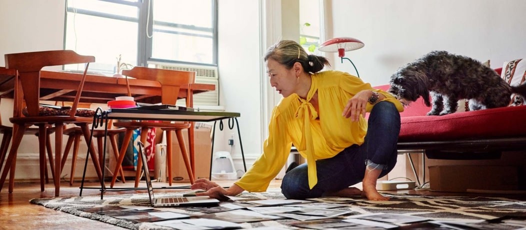 Asain woman in a bright yellow blouse and jeans on sitting on her living room floor on her laptop. A dog is on the couch behind her.