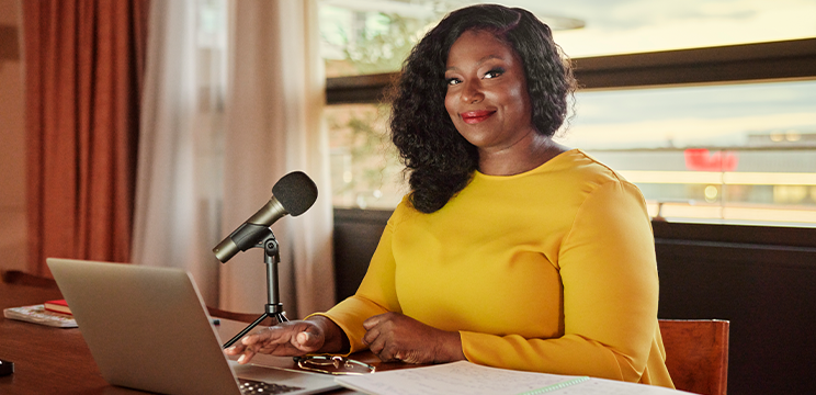 Woman seated at a desk with a laptop and microphone. She is smiling.