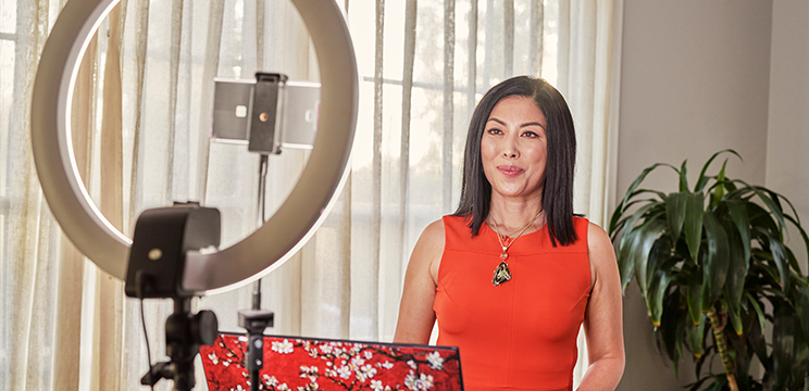 A woman in a red dress takes a photo of herself using her phone on a stand with a ring light.