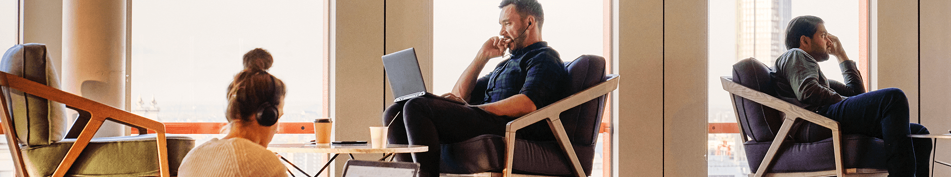 Three individuals sitting in a cafe a women on the left on her laptop, a man in the middle intently focusing in on his laptop, and a man on the right on a phone call