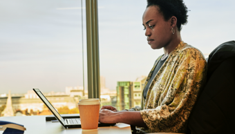 Femme en pleine réflexion, travaillant sur son ordinateur dans un bureau avec une vue sur la ville.