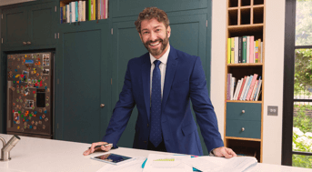 Man in suit stands behind table in front of bookshelf