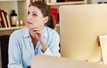photograph of a woman staring off to the left side of the screen while sitting at a desk with a large monitor