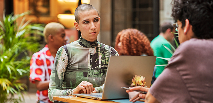 Two people sit opposite one another at an outdoor table. One has a laptop open and is listening intently.