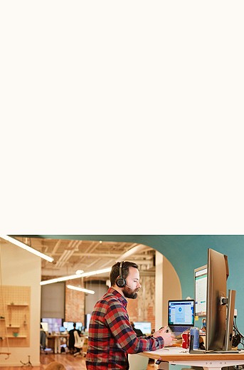 photograph of a male with a headset at a standing desk, working on three large monitors