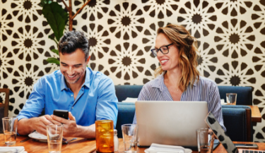 Two business people laugh and work together at a cafe table, with a phone and computer