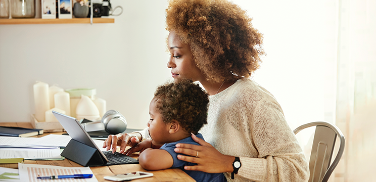 A woman works at her laptop with her toddler in her lap.
