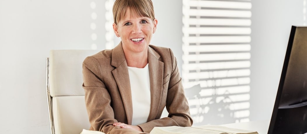 Caucasian woman in a brown blazer sitting at her desk looking at the camera.