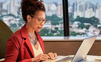 Photograph of a woman with her hair up wearing red glasses and a red blazer working on a laptop computer at a desk near a window.
