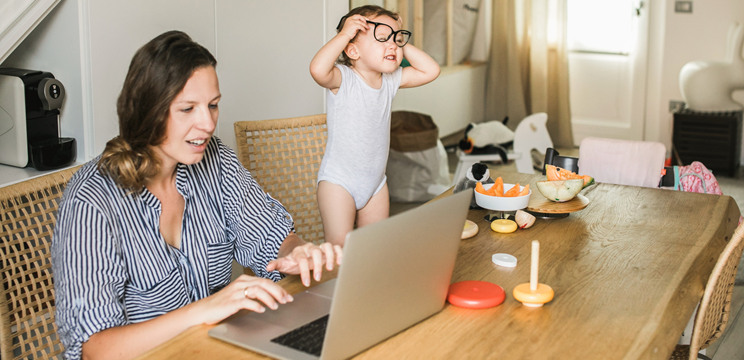 An image of a woman working at a laptop while her toddler tries on her glasses.