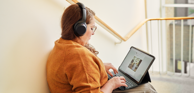 A woman sitting on a stairwell, typing on her laptop while wearing headphones.