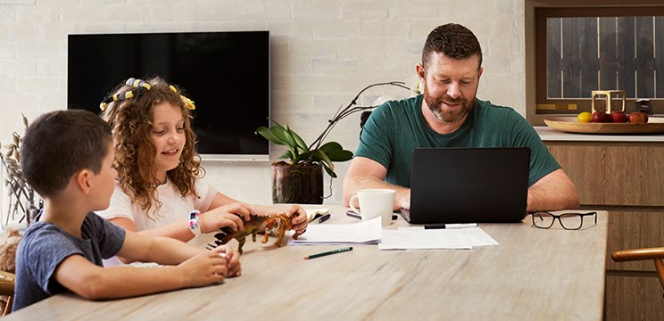A man works on his laptop at home while his children play beside him.