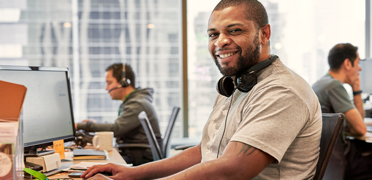 A person sitting at a desk with headphones around their neck smiles at the camera.