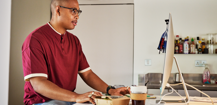 Image of a man with a red shirt sitting at his work desk and staring at his Mac desktop screen while he is working.