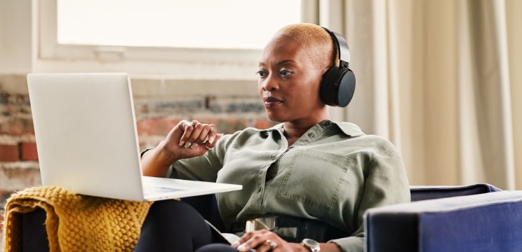 A woman wearing headphones watches something on her laptop while sitting in an armchair.
