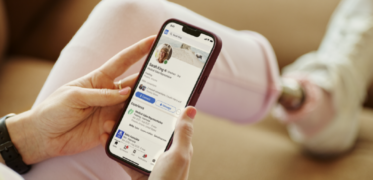 Image of a person’s hands holding their phone. They have the LinkedIn app open and are looking at another member’s profile.