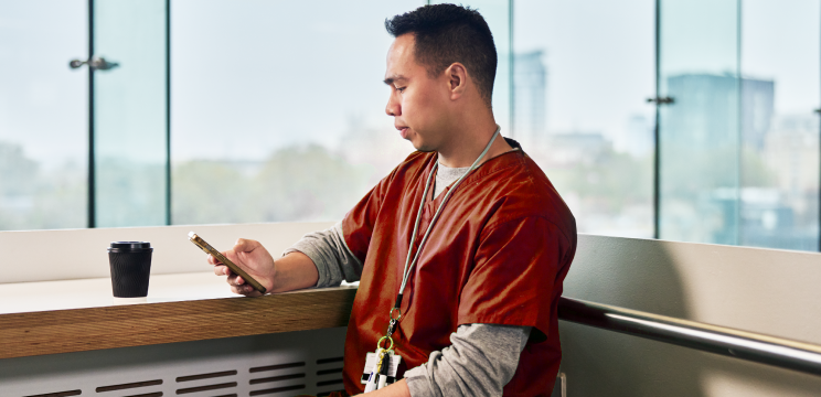 Male healthcare worker is seated in an office setting looking at something on his phone with a black coffee cup on the table in front of him.