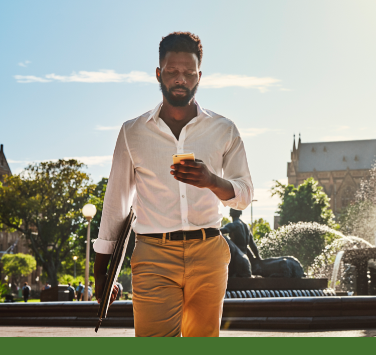 Man walking outdoors looking at his phone with a laptop under his arm