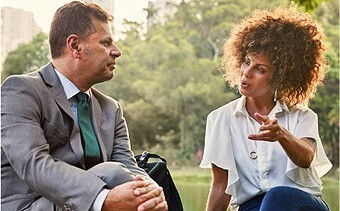 Photograph of a woman and a man sitting outside in a city park. The woman is talking and wearing a short-sleeved white blouse and the man is sitting cross-legged wearing a grey suit with forest green tie.