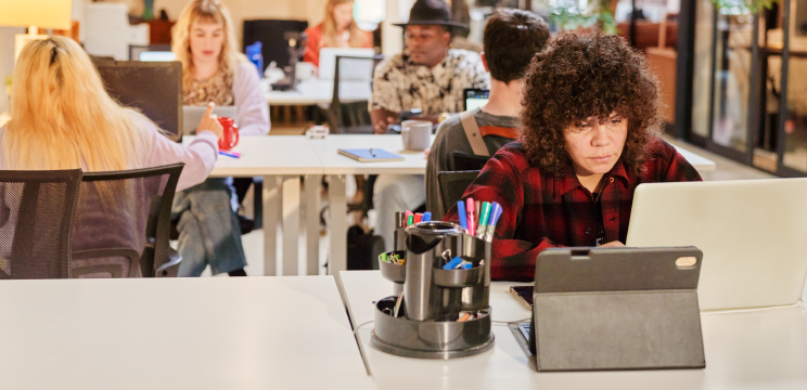 A person works on their laptop in an open-plan office. A tablet and stationery holder are on the desk beside them.