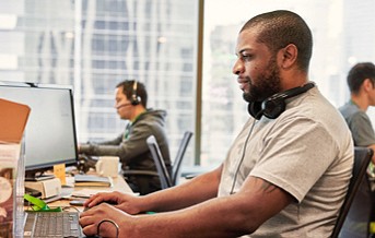 photograph of a man working on a laptop in an open-office concept with headphones sitting on shoulders, with another man in the background