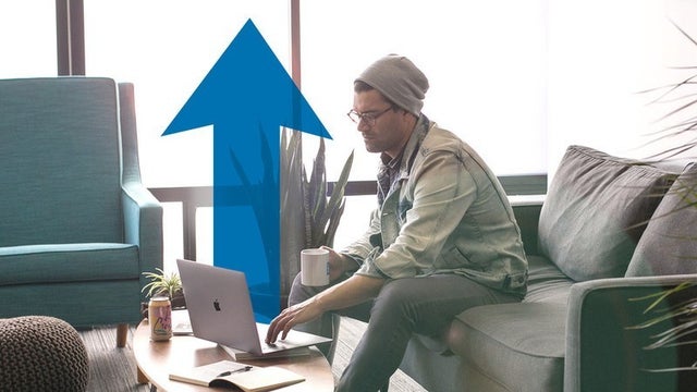 A man sits on a couch working on his computer, with an illustrative blue up arrow behind him