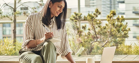 Image of a woman looking at her laptop while writing notes on her notepad