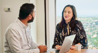 Two people sit at a table talking in front of a laptop