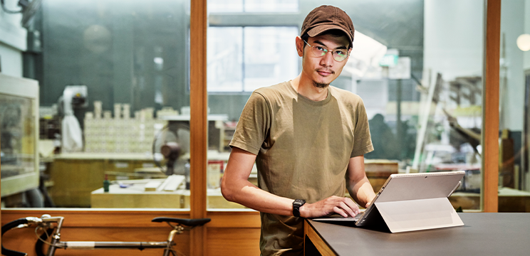 A person looks at the camera while standing at a desk with a laptop open in front of them.