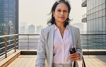 Photograph of a professional looking woman standing on a rooftop holding a purse on her wrist and a cellphone in her left hand