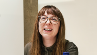 Femme avec des lunettes assise derrière son ordinateur et riant avec quelqu’un hors cadre.