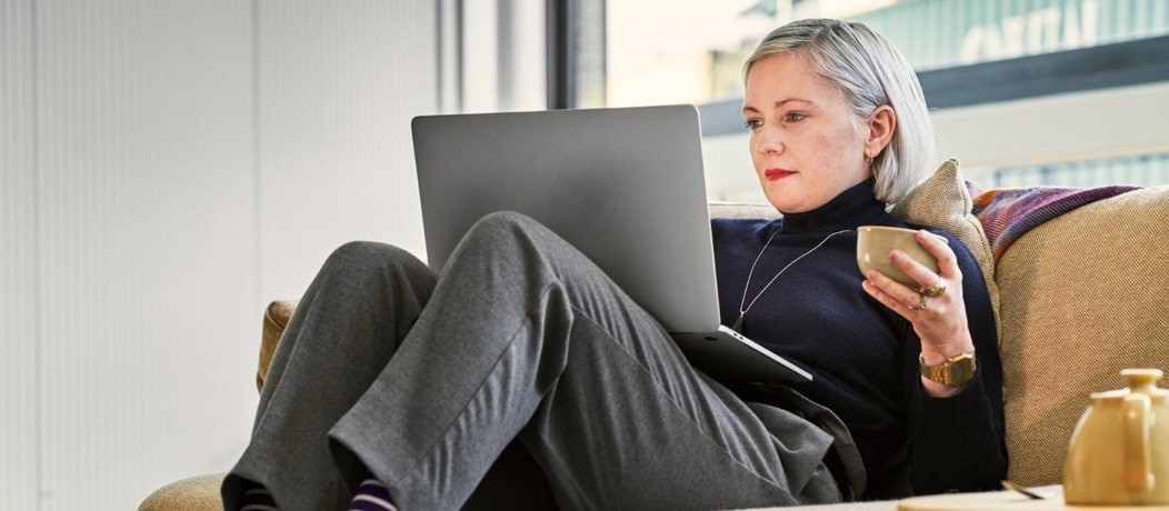 Grey haired Caucasian woman sitting on her couch with a cup of tea and her laptop.