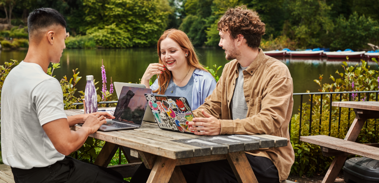 Three people working together at an outdoor picnic table with laptops beside a lake, illustrating collaborative volunteer engagement.