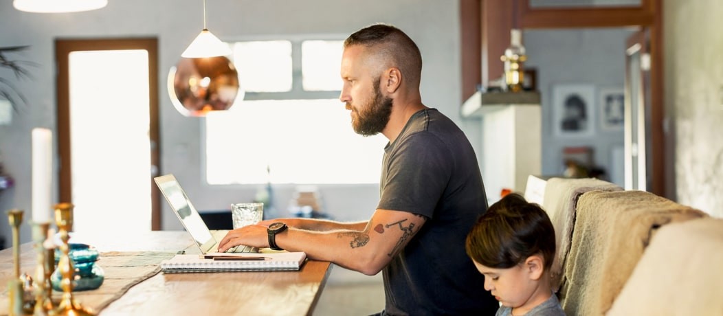 Caucasian man with a beard and arm tattoos sitting at a table with a laptop and his son next to him.