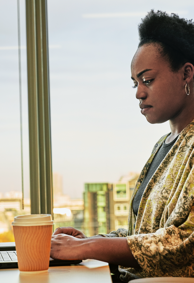 Femme en pleine réflexion, travaillant sur son ordinateur dans un bureau avec une vue sur la ville.