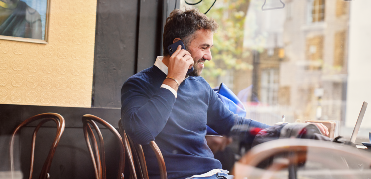 A person sitting in a cafe taking a phone call while using their laptop. They are smiling.
