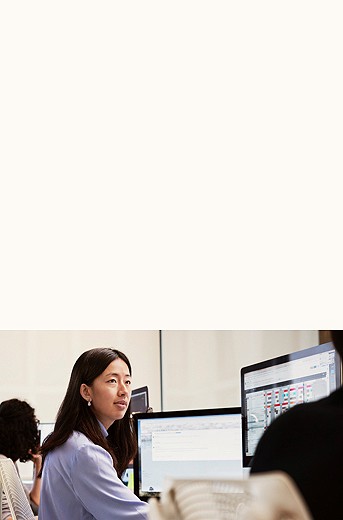 Photograph of a woman looking over to someone off screen sitting at a desk with two computer monitors
