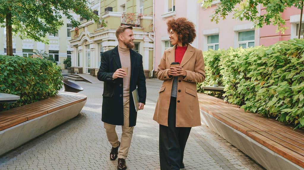 A man and a woman in coats walking and talking together along a tree-lined city street with coffee in hand