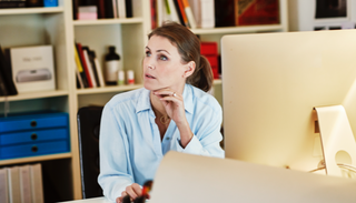 Femme avec le menton posé sur la main, assise à un bureau devant un ordinateur dans une entreprise. Elle regarde et écoute une personne en dehors du cadre. Une grande étagère de livres se trouve en arrière-plan.