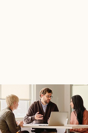 Photograph of a man and two women sitting together at a round table talking over one single laptop.